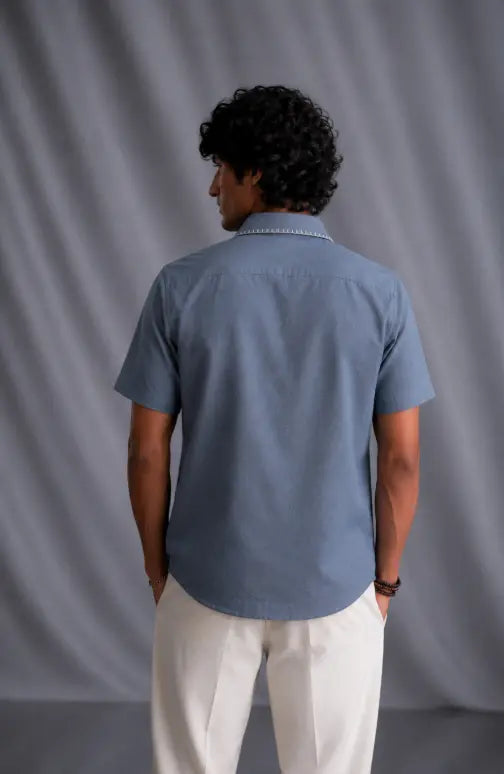 A man viewed from behind wearing a light blue short-sleeve button-up shirt and off-white trousers, standing against a gray draped studio backdrop.