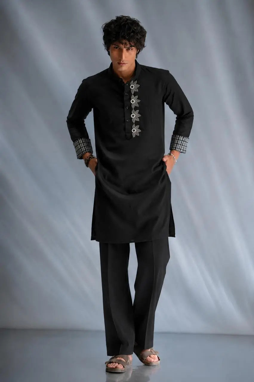 A man modeling a black long-sleeve kurta with decorative silver-toned buttons down the front, matching black trousers, and checkered cuffs, posed against a gray draped studio backdrop.