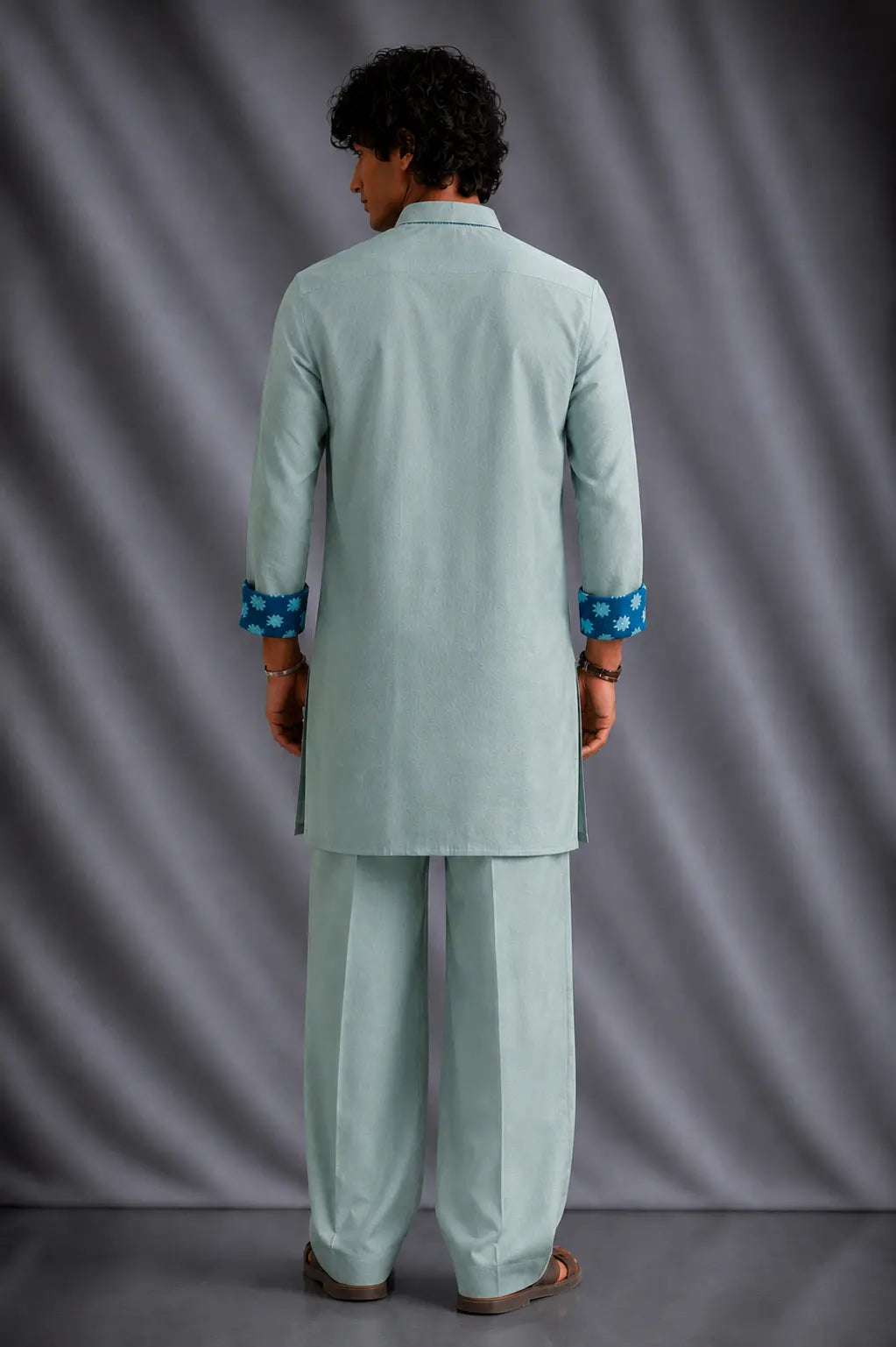 A man viewed from the back wearing a light sage green kurta and matching trousers, featuring blue patterned cuffs, set against a gray draped studio backdrop.