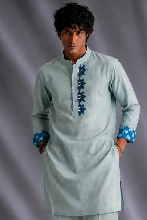 A man modeling a light sage green kurta and matching trousers set, featuring blue patterned button details on the chest and cuffs, set against a gray draped studio backdrop.
