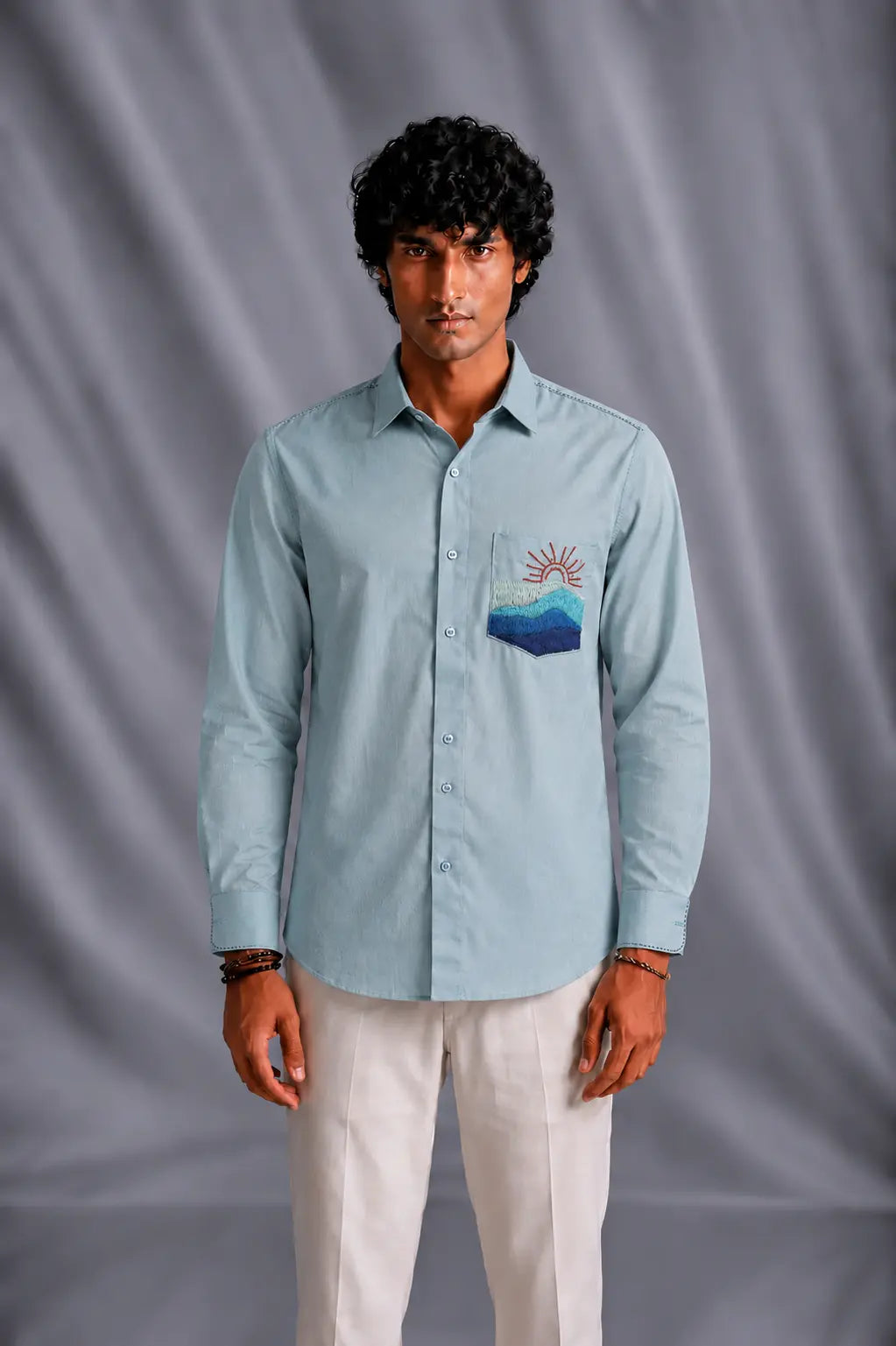 A man wearing a light blue long-sleeve button-up shirt with a stylized mountain and sun patch on the chest pocket, paired with off-white trousers, standing against a gray draped studio backdrop.