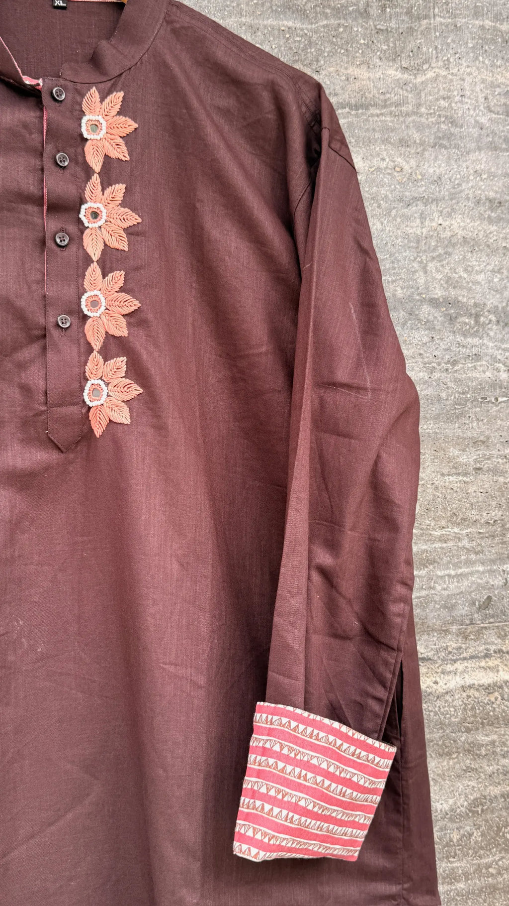 A close-up of a brown long-sleeve kurta or tunic with embroidered floral details on the button placket and a striped patterned cuff, shown against a gray textured background.