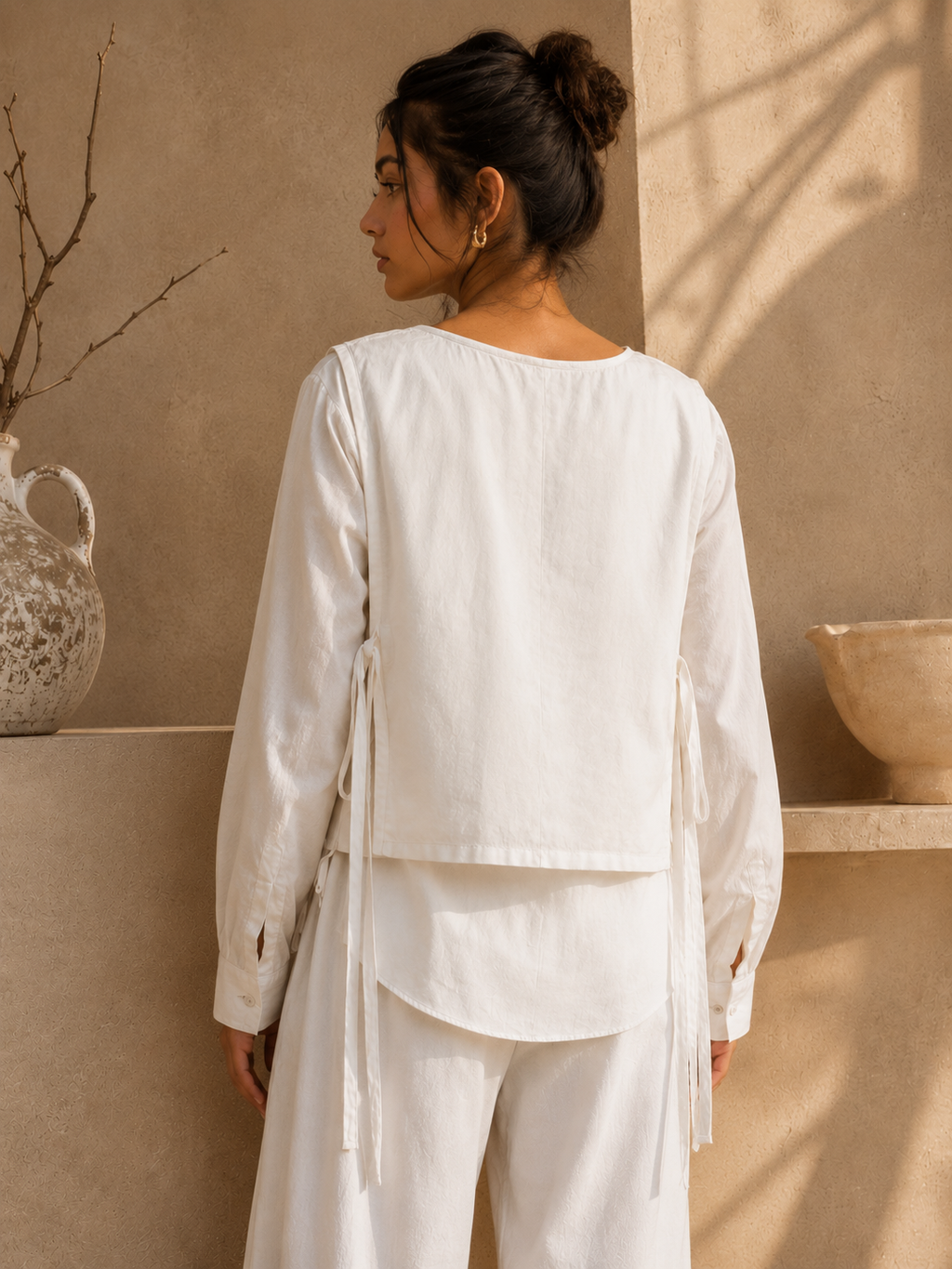 A woman shown from the back wearing a white long-sleeve top and matching white pants in a warm beige interior setting with decorative pottery and soft shadows.