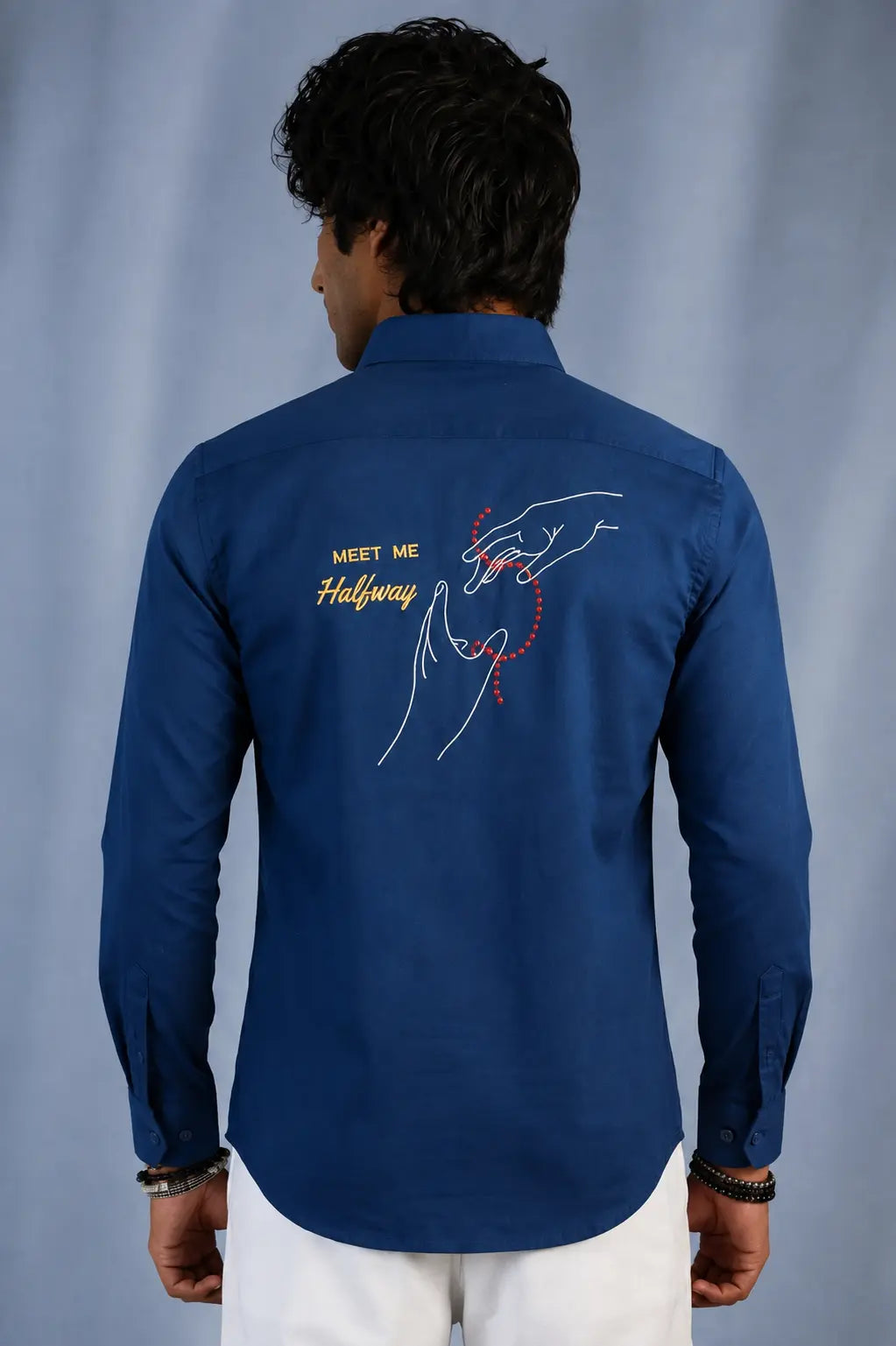 A man viewed from the back wearing a royal blue long-sleeve shirt with a graphic print on the back. The print features a white line-art illustration of a hand holding a red heart, alongside the text 'MEET ME Halfway' in yellow and white.