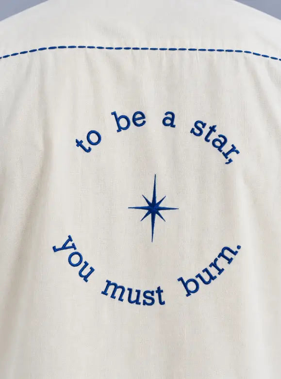 Close-up of a white fabric garment with blue embroidery, featuring the text 'to be a star, you must burn.' arranged in an arc around a blue starburst motif.